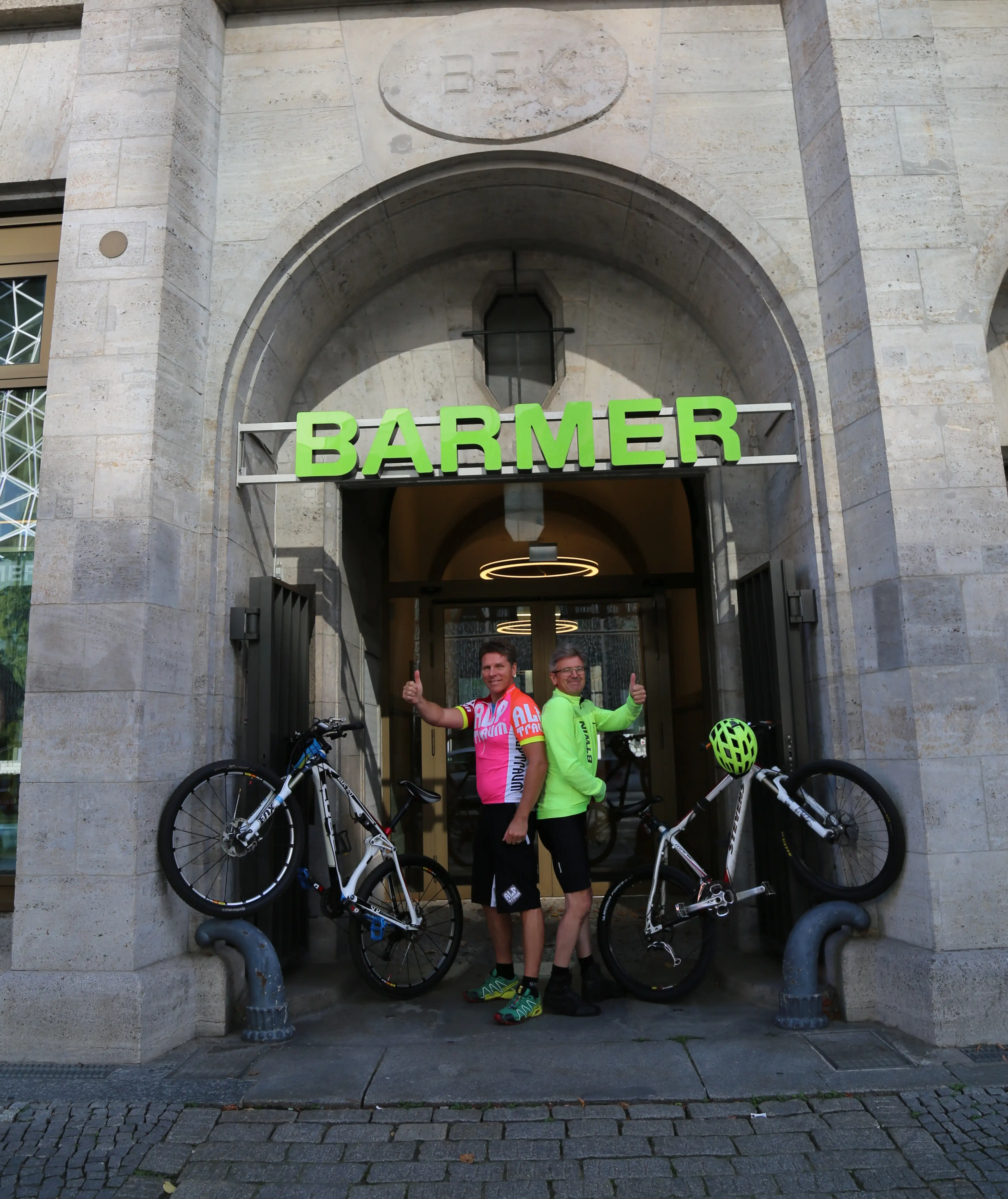 Zwei Barmer Mitarbeiter, die mit ihren Fahrräder ein V in der Eingangspforte von Barmer bilden Zwei Barmer Mitarbeiter, die mit ihren Fahrräder ein V in der Eingangspforte von Barmer bilden